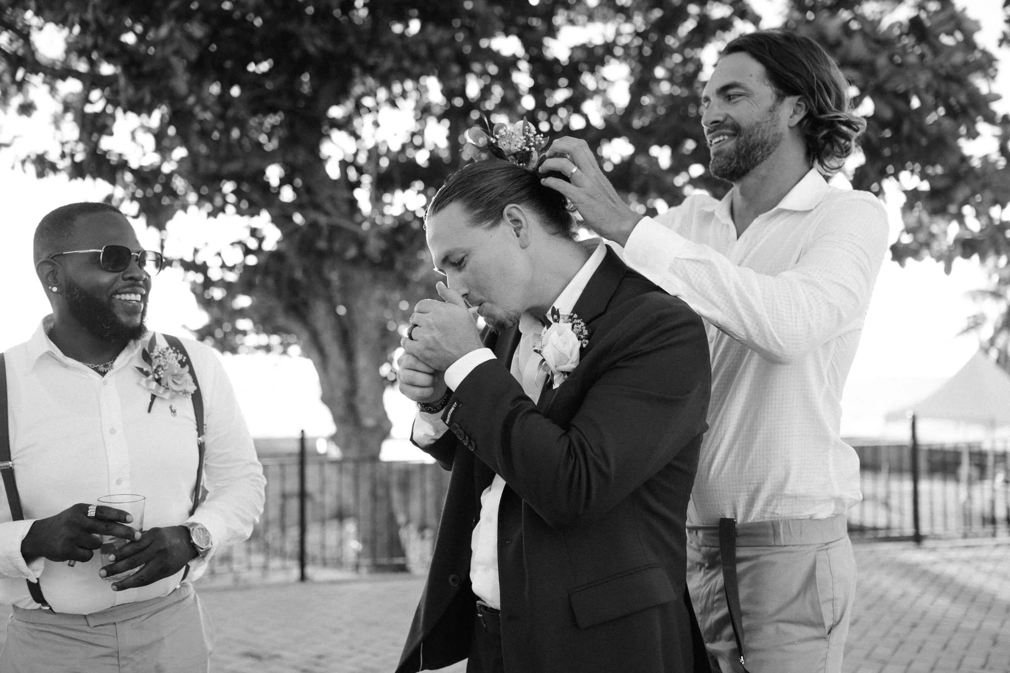 A black-and-white photo of a wedding ceremony outdoors, showing a groom in a suit praying, a person placing a floral crown on his head, and another man smiling.
