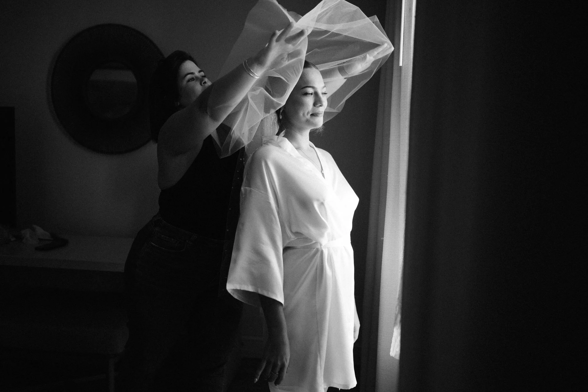A woman in a satin robe stands next to a window as a stylist helps her put on a sheer veil.