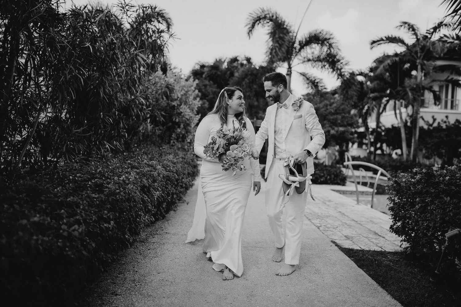 A black and white photo of a bride and groom walking barefoot on a path outdoors, holding hands and smiling at each other. The bride is holding a bouquet of flowers, and the groom is carrying a pair of shoes and a hat.