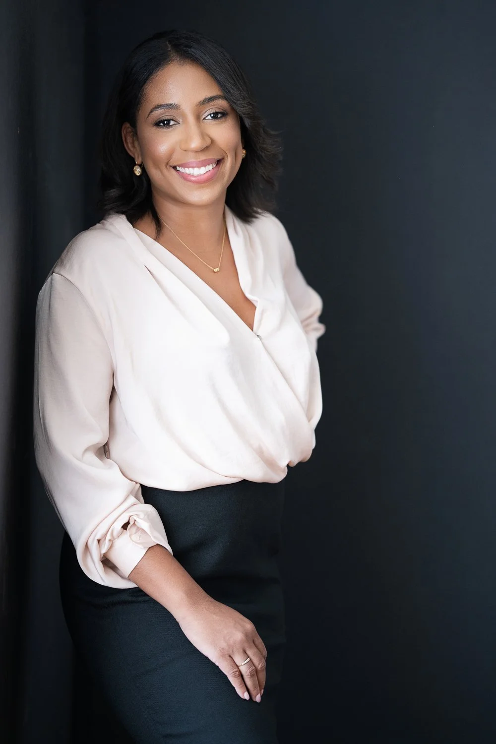 A woman with dark hair, wearing a light-colored silky blouse and black skirt, smiling and standing against a dark wall.