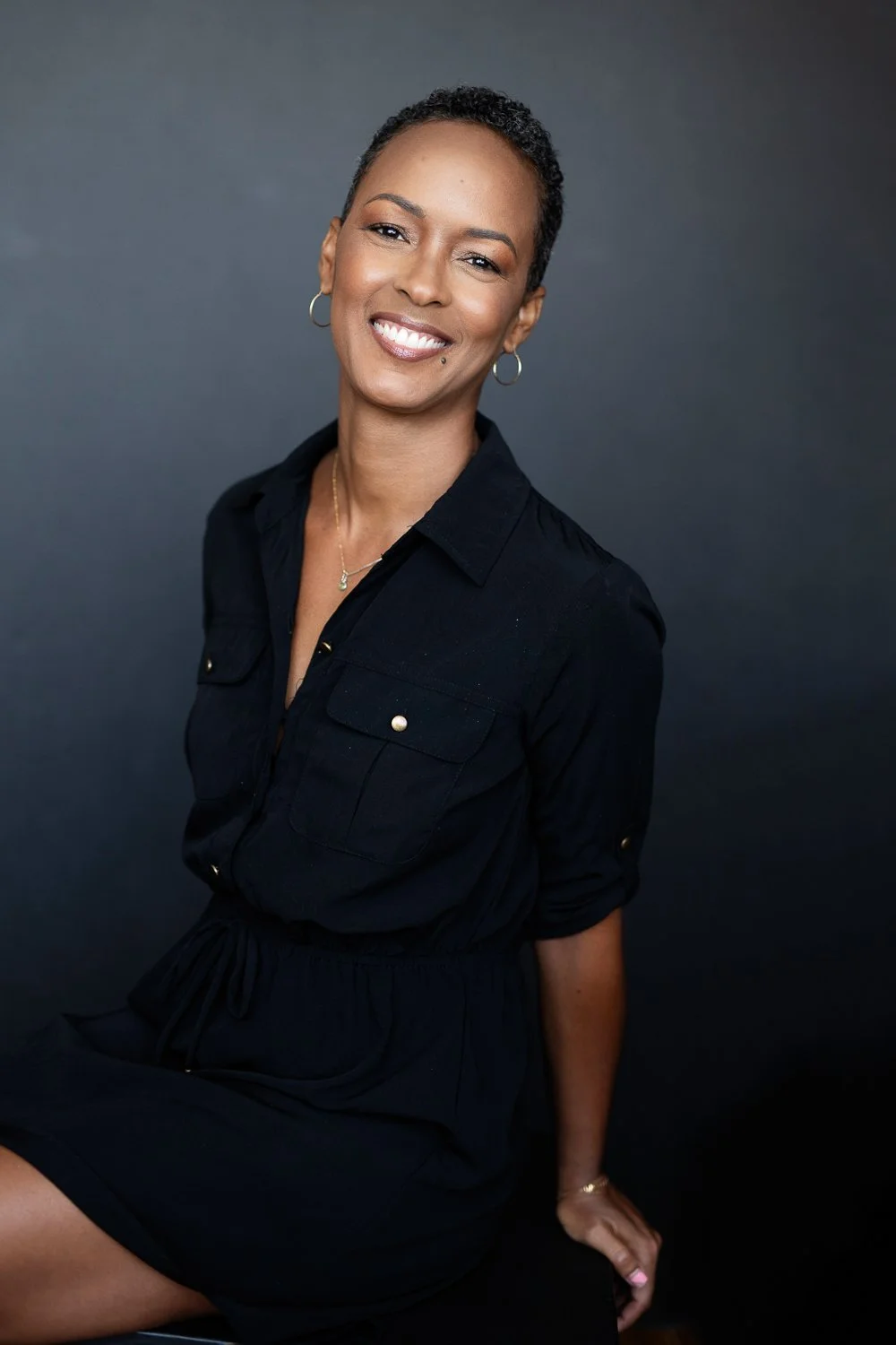 A woman with short curly hair, smiling, wearing a black dress with a collar and pocket details, silver hoop earrings, and a delicate necklace, sitting against a dark gray background.