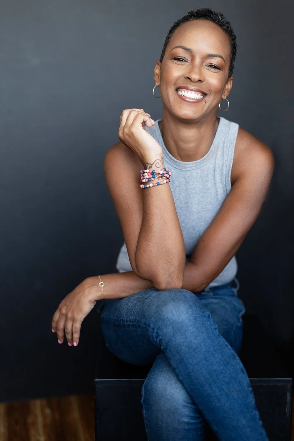 A woman with short curly hair smiling and sitting against a dark background, wearing a gray sleeveless top, ripped jeans, earrings, colorful beaded bracelets, and a small nose ring.