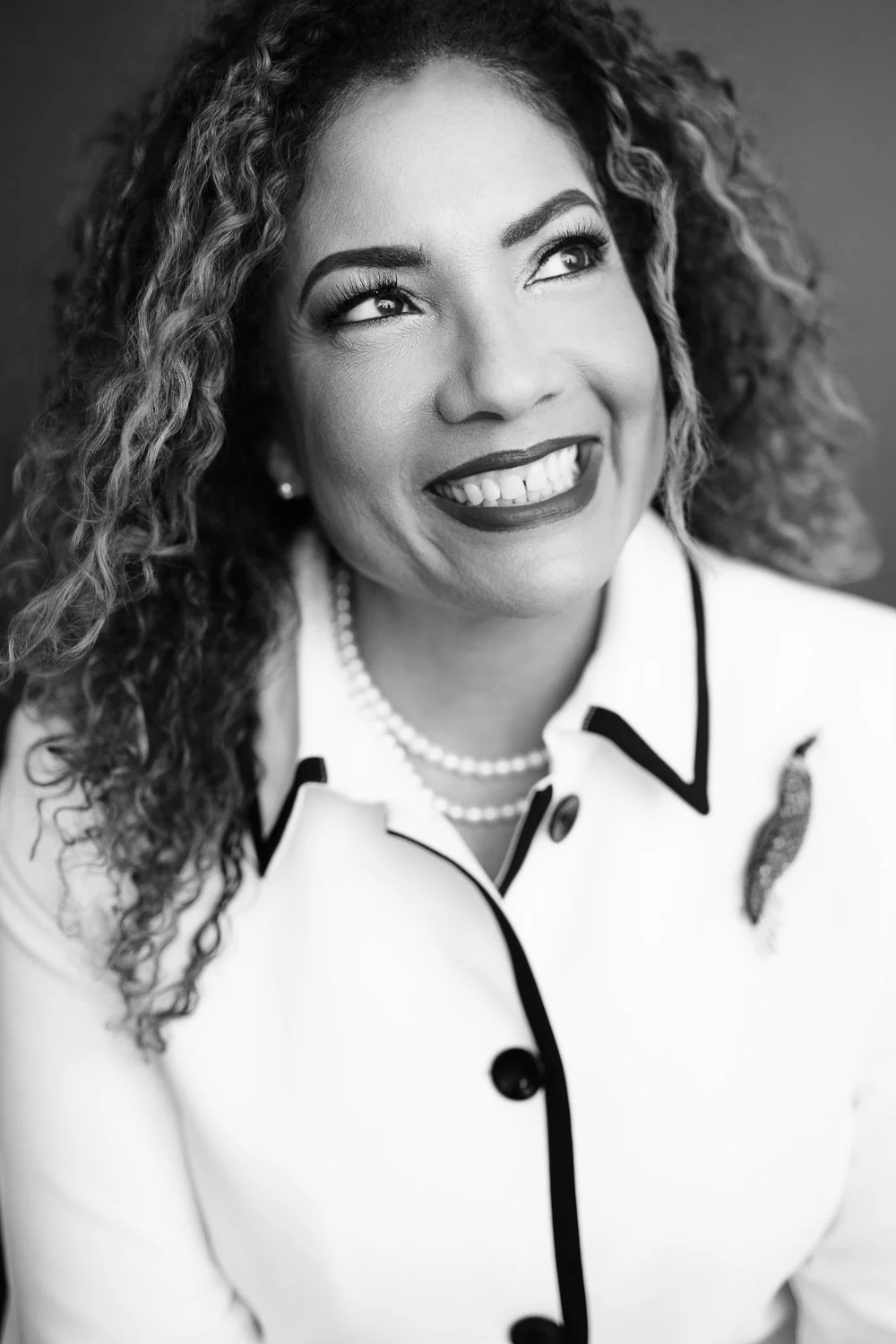 Black and white portrait of a woman smiling, wearing a blazer with a leaf-shaped pin, pearl necklaces, and earrings, with curly hair and bright eyes.